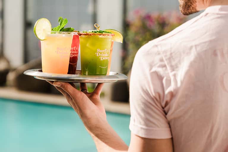 Server holding tray of colorful cocktails labeled 'Surfly Drink Down' by resort pool