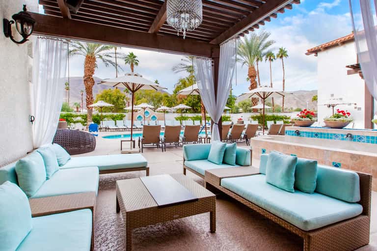 Elegant covered lounge area with turquoise cushioned seating, pool views, and palm trees in desert landscape
