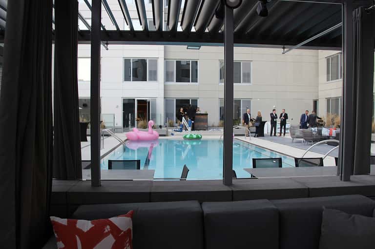 Indoor pool viewed through floor-to-ceiling windows with black pergola and people in background