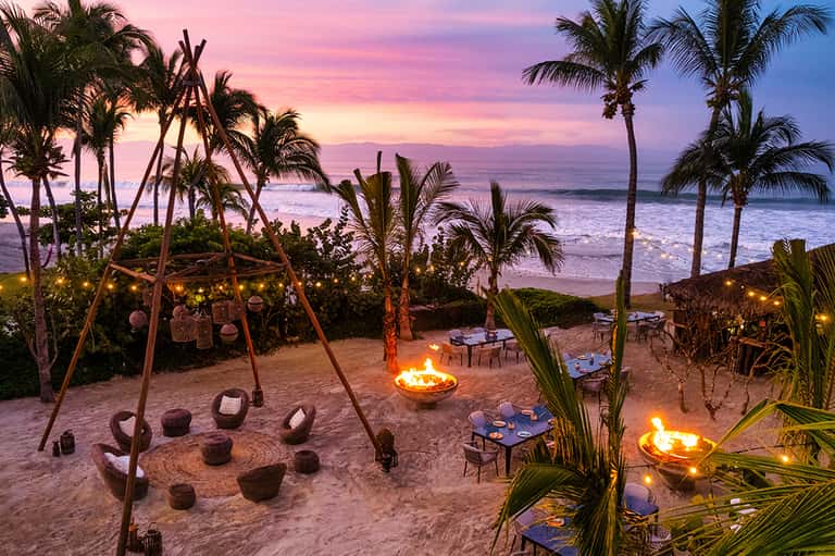 Beach bonfire at sunset with palm trees, lounge seating, and pink-purple sky over ocean