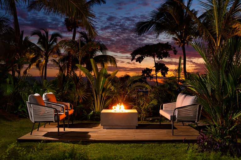 Beachfront fire pit seating area at dusk with lounge chairs, palm trees, and dramatic sunset sky