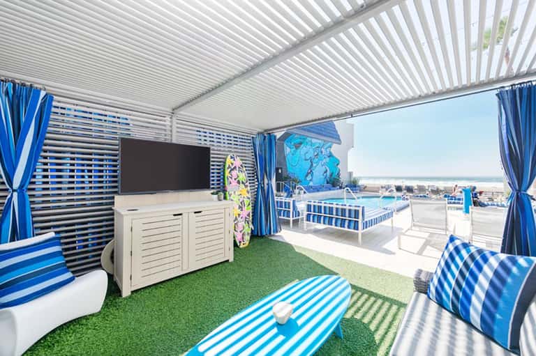 Covered beach cabana with blue striped furnishings, TV, and ocean view beyond railing
