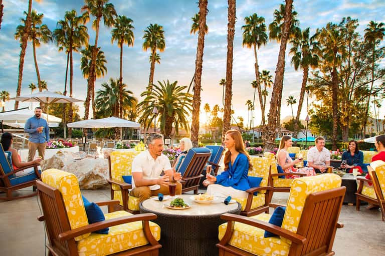 Outdoor seating area with guests in yellow and blue chairs, palm trees, and mountains