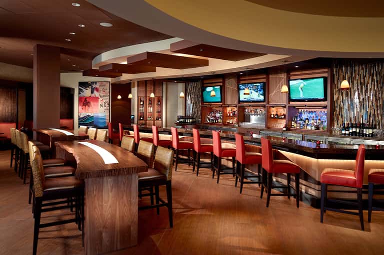 Upscale resort bar lounge with dark wood, red chairs, multiple TV screens, and curved ceiling