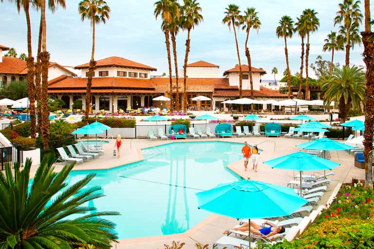 Sprawling resort pool complex with turquoise water, blue umbrellas, palm trees, and Spanish-style architecture