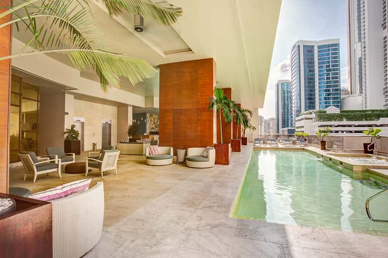 Modern rooftop pool area with turquoise water, lounge seating, tropical plants, and city skyline views