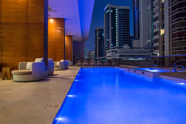 Rooftop infinity pool at dusk with glowing blue water, lounge seating, and city skyline backdrop