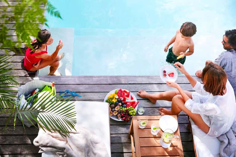 Family enjoying poolside lunch and drinks on wooden deck with tropical flowers and blue water
