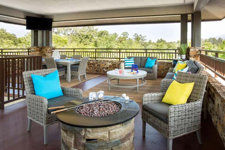 Elevated deck lounge with wicker furniture, fire pit, colorful pillows, and forest views