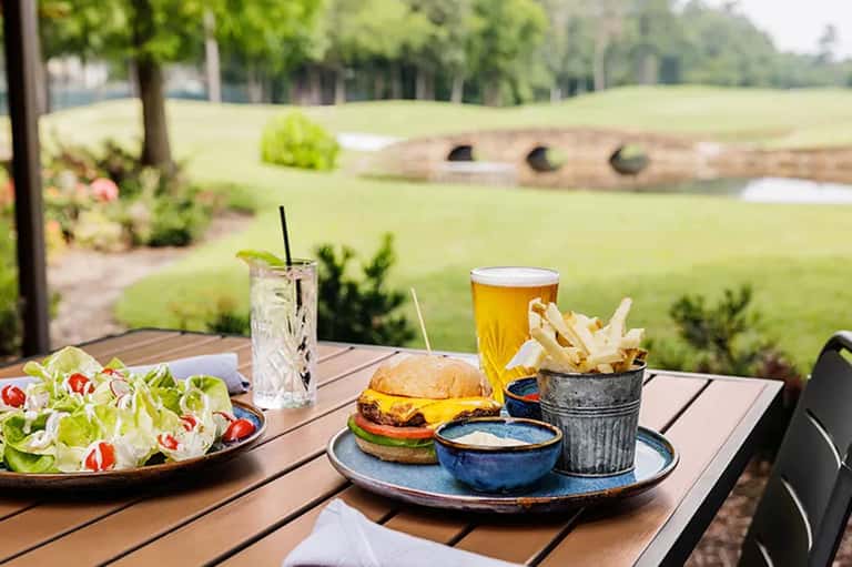 Outdoor dining table with burger, fries, salad, and beverages overlooking golf course with tree-lined fairway and water hazard