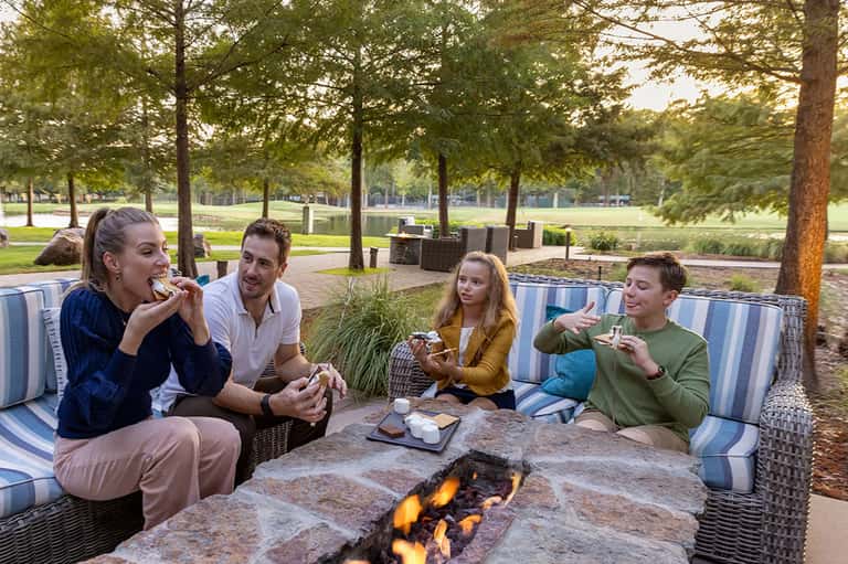 Family gathered around outdoor fire pit with food, striped cushioned seating, and lakeside trees