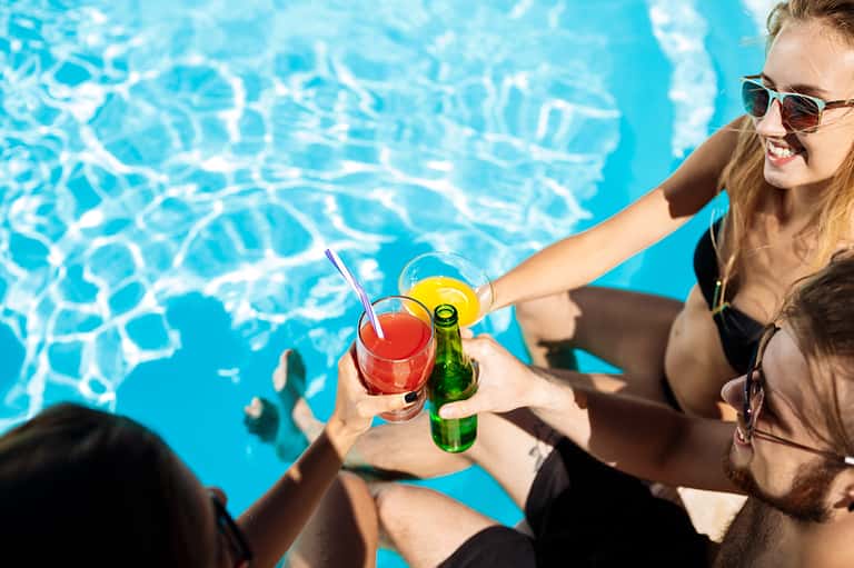Woman at pool holding colorful cocktail and beer bottle, smiling with clear blue water background