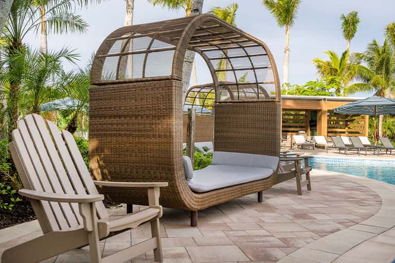 Woven rattan lounge pod with gray cushion by resort pool, flanked by Adirondack chair and palm trees