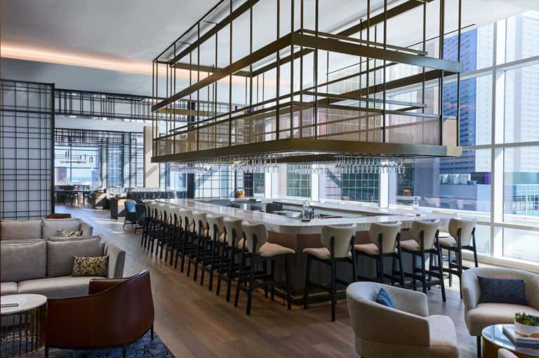 High-rise hotel bar with floor-to-ceiling windows, industrial metal ceiling framework, and city views