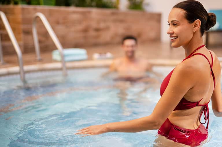 Woman in red swimsuit in a resort pool smiling, with other guests visible in background and brick walls