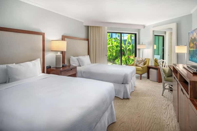 Hotel guest room with two queen beds, wooden headboards, green accent chair, glass doors to patio, and warm lighting
