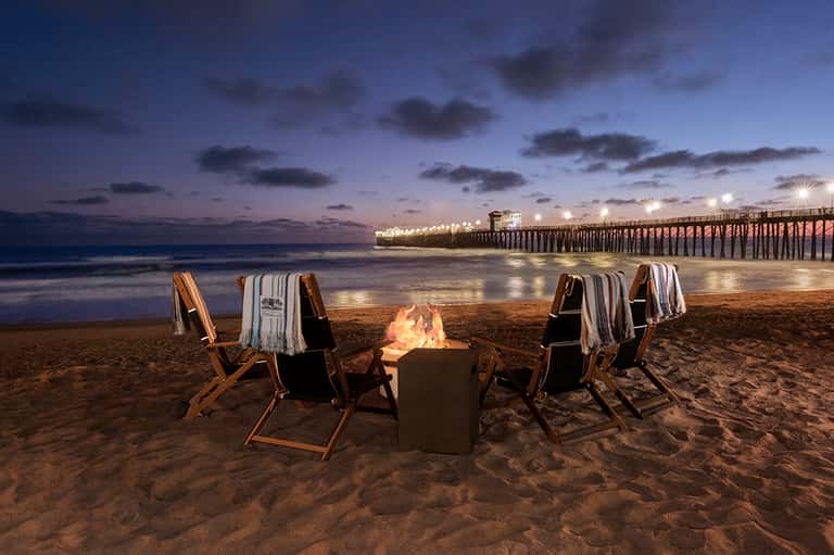 Beach bonfire at dusk with lounge chairs, blankets, illuminated pier, and ocean view at twilight