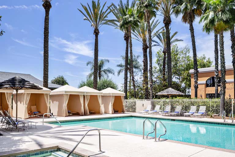 Resort pool with turquoise water, white cabanas with curtains, palm trees, and lounge chairs on sunny day