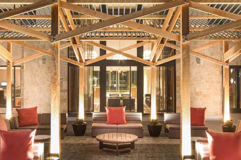 Covered outdoor lounge with dramatic wooden beams, gray seating, red pillows, and ambient lighting