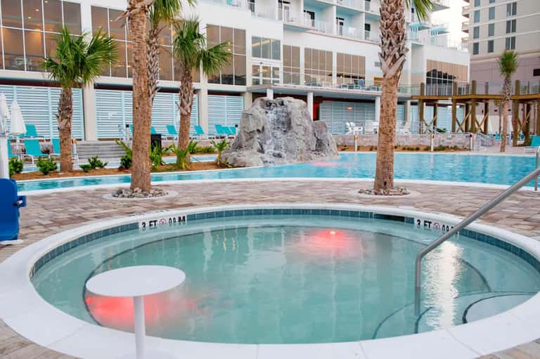 Hot tub and main pool area with palm trees, modern hotel building, and beach cabanas