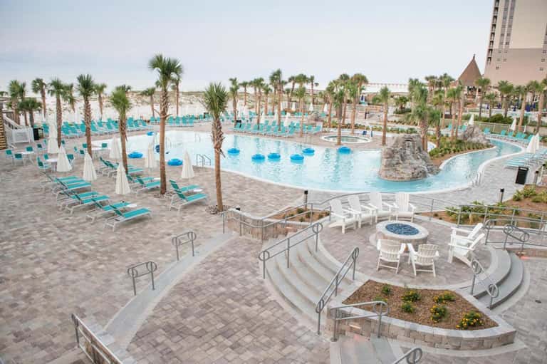 Large resort pool complex with turquoise water, palm trees, lounge chairs, and high-rise hotel building