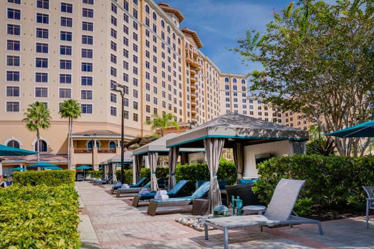 Resort pool deck with lounge chairs, cabanas, palm trees, and multi-story hotel building