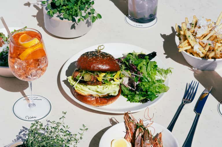 Outdoor dining table with gourmet burger, fries, and cocktail on sunny terrace with potted plants