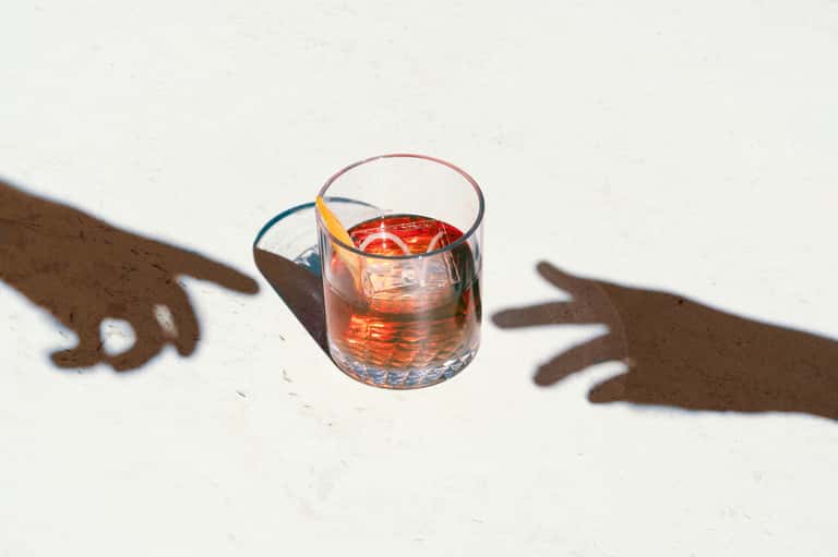 Glass of amber cocktail with shadow of hands on white surface in sunlight