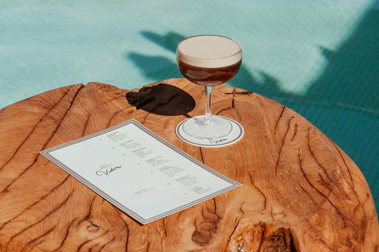 Elegant wooden table with cocktail menu and espresso martini overlooking turquoise pool waters