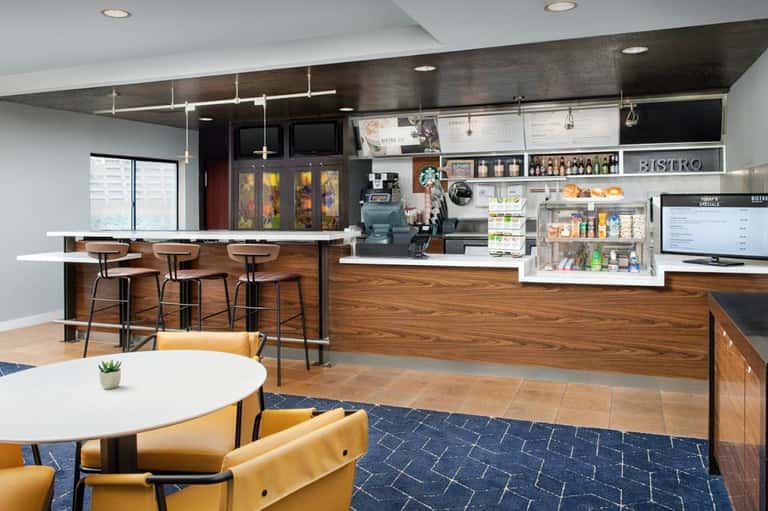 Modern hotel bistro with wood counters, bar seating, white shelving, and casual dining tables with yellow chairs