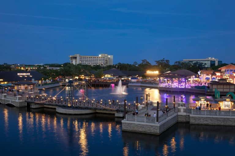 Illuminated waterfront entertainment complex at dusk with fountain, bridges, and colorful purple-lit buildings