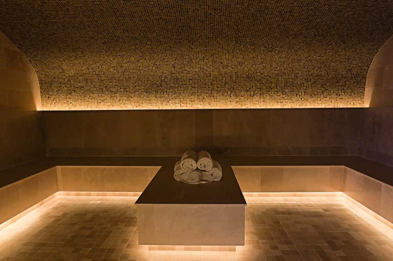 Luxurious spa treatment room with mosaic tile ceiling, heated stone basin, and rolled towels on dark stone platform