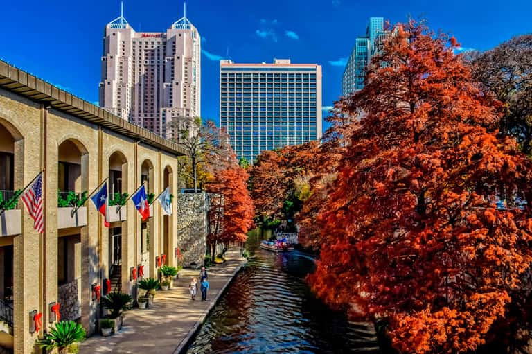 Scenic waterfront promenade with historic stone building, modern skyscrapers, and vibrant red autumn foliage