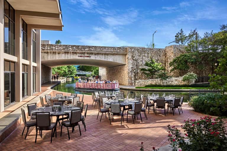 Waterfront terrace dining area with round tables, chairs, and canal view under modern building overhang