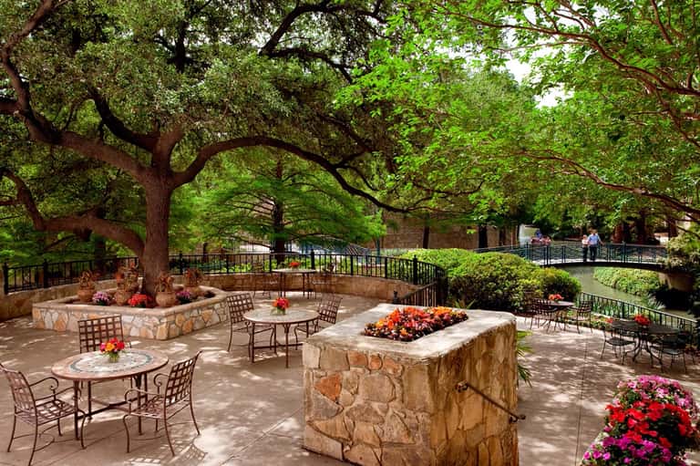 Shaded outdoor patio with stone seating areas, flower beds, and metal railings overlooking a garden pathway
