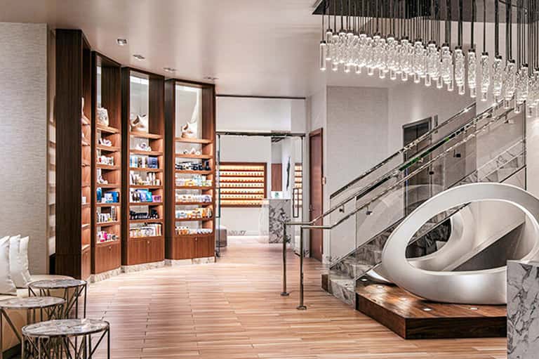 Modern luxury spa reception area with illuminated shelving, marble counters, and contemporary staircase with hanging glass chandelier