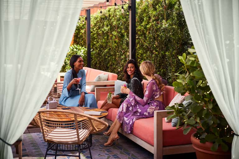 Three women socializing on coral pink seating under white curtains in an elegant garden patio setting