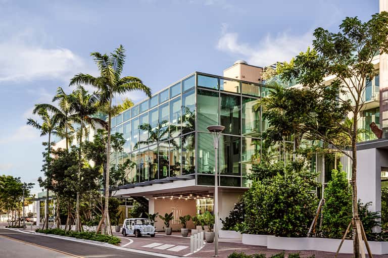 Modern glass-walled building with green-tinted windows surrounded by palm trees and tropical landscaping on a street corner.