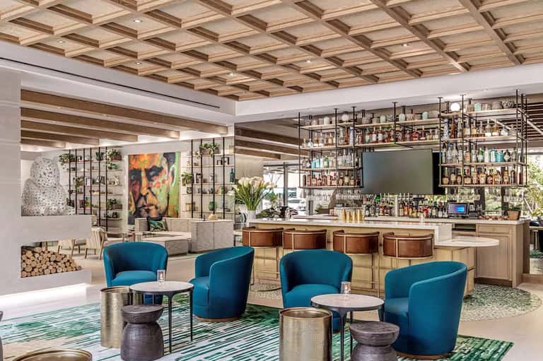 Modern hotel lounge with coffered wood ceiling, teal armchairs, and full-service bar with extensive bottle selection