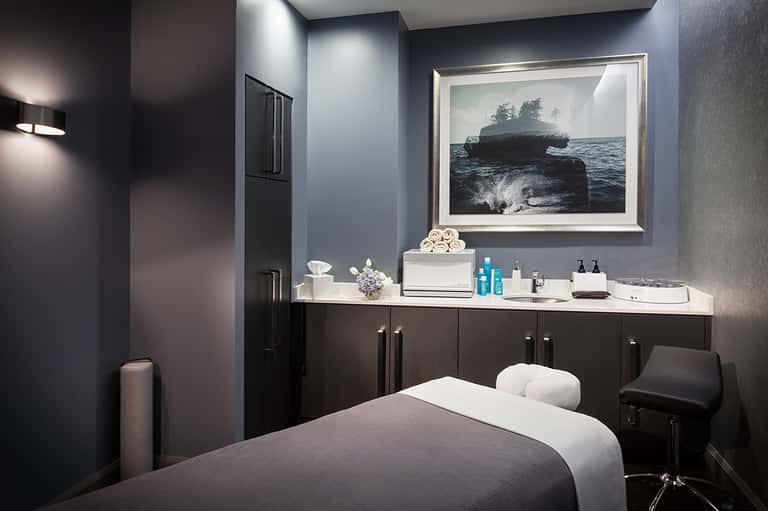 Modern spa treatment room with dark cabinetry, white massage bed, and ocean landscape artwork on blue wall