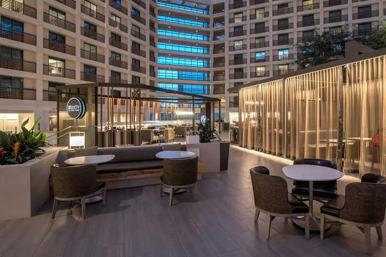 Modern hotel courtyard with lounge seating, round tables, and illuminated glass partitions under residential towers