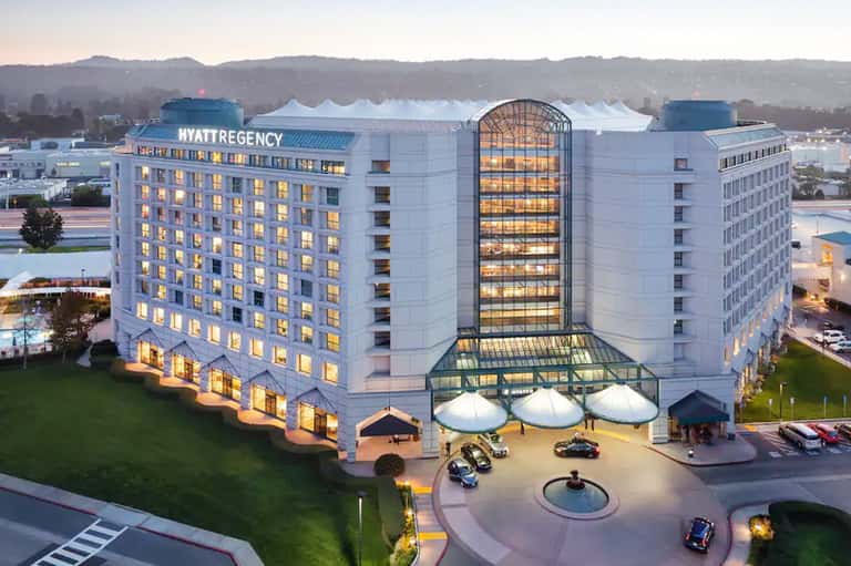 Hyatt Regency hotel at dusk with illuminated rooms, white tent structures in courtyard, mountains in background