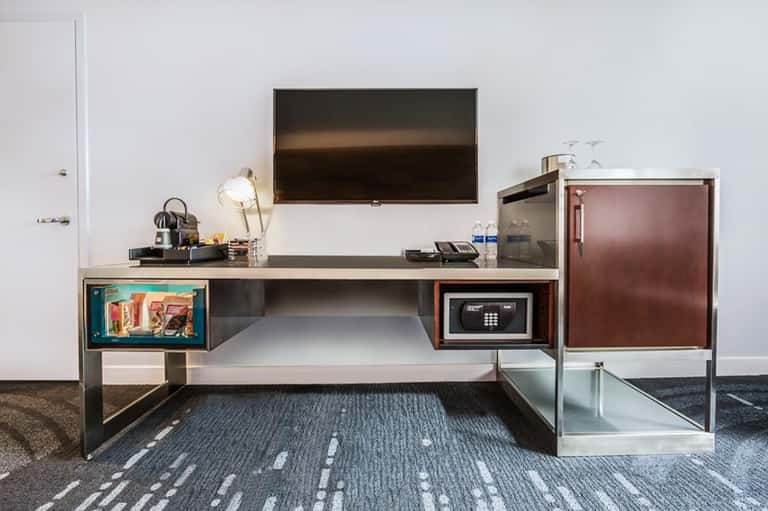 Modern hotel room desk with TV, safe, minibar, and work lamp on textured carpet flooring