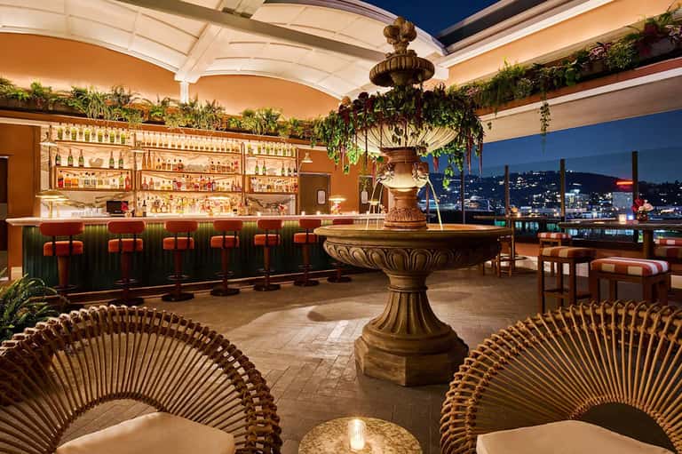 Upscale rooftop bar with golden fountain, woven chairs, city skyline views, and illuminated liquor shelves