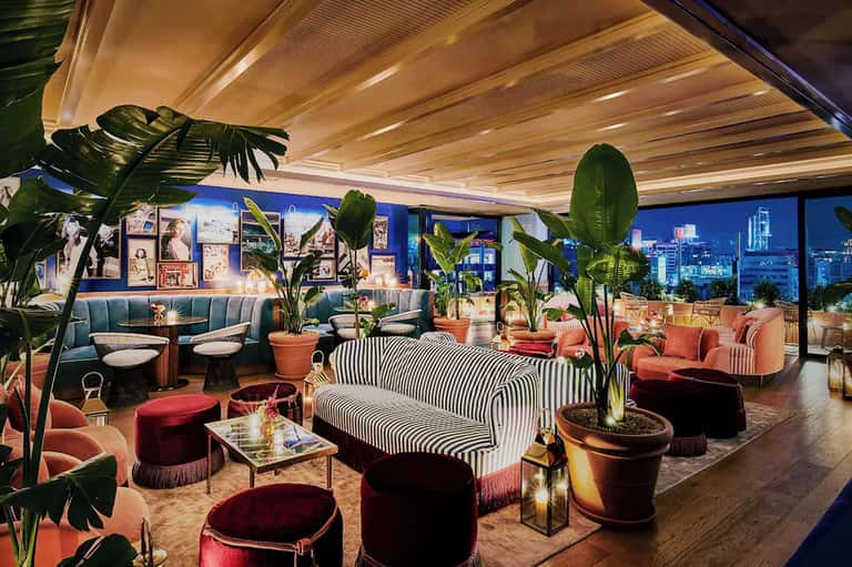 Luxury lounge with blue and coral seating, striped sofa, potted plants, wooden ceiling, and city skyline views at night