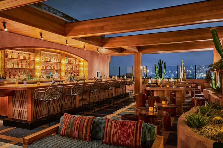 Luxury rooftop bar at twilight with cityscape views, wooden beams, illuminated liquor display, and modern seating