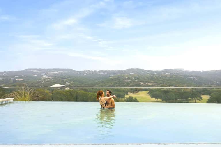 Couple relaxing in infinity pool overlooking lush green hillside landscape and valley views