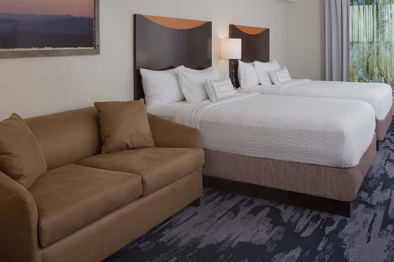 Modern hotel room with two queen beds, white bedding, tan sofa, and patterned carpet flooring