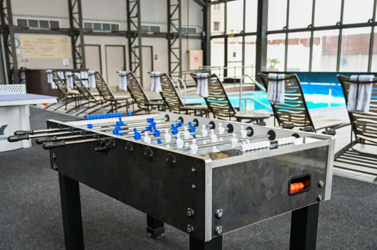 Foosball table by pool with lounge chairs and large windows overlooking waterfront