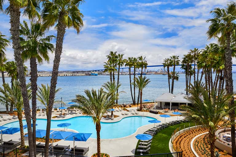 Beachfront resort with curved blue swimming pool, palm trees, and ocean view with ships in harbor